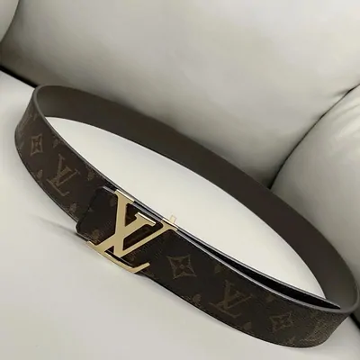 A brown monogram-patterned leather belt coiled on a cream sofa, fastened with a large gold-tone rectangular buckle.