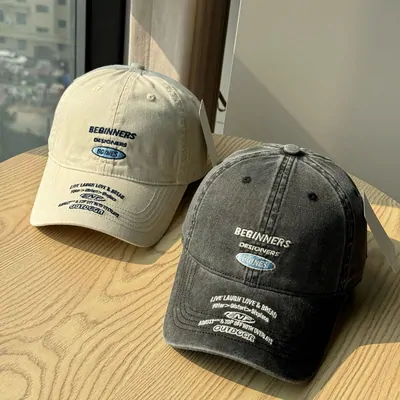 Two baseball caps on a wooden table, one in washed cream and one in faded charcoal, both with small embroidered lettering on the front panel.