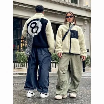 Two people standing on a city pavement in oversized fleece jackets, one cream with a dark circular panel on the back, worn with wide-leg trousers.