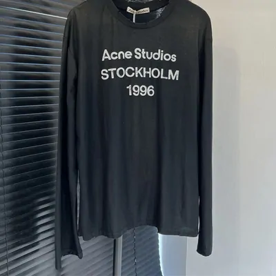 A black long-sleeve cotton tee on a wire hanger against a pale wall, with three lines of faded white lettering across the chest.