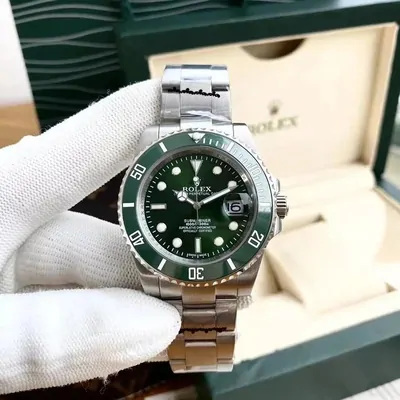 A gloved hand holding a stainless steel dive watch with a green dial and a green bezel, its open presentation box behind.